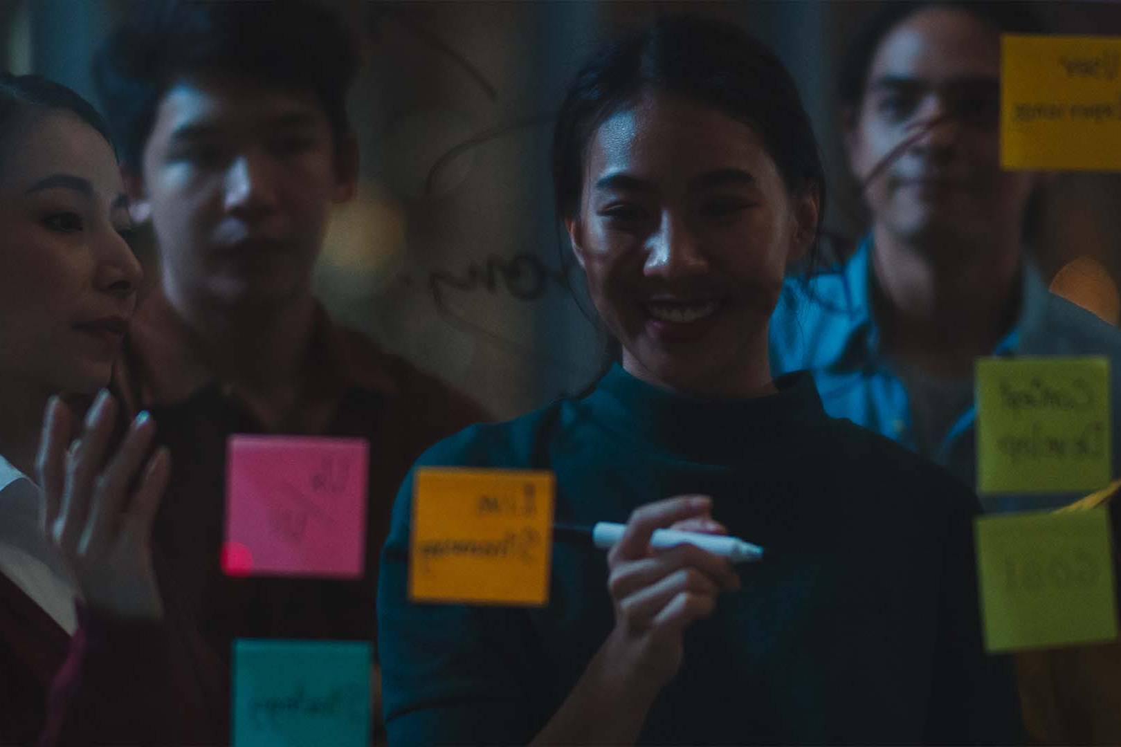 A smiling female professional writing on a glass board covered in colorful sticky notes while her team watches.