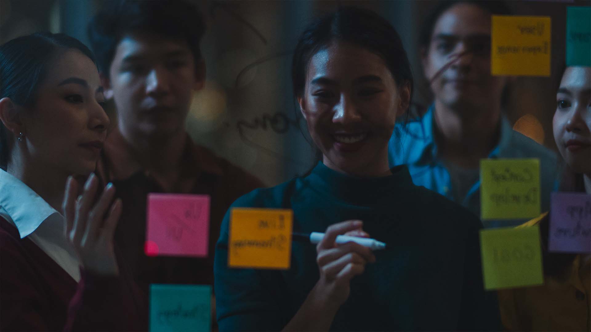 A smiling female professional writing on a glass board covered in colorful sticky notes while her team watches.