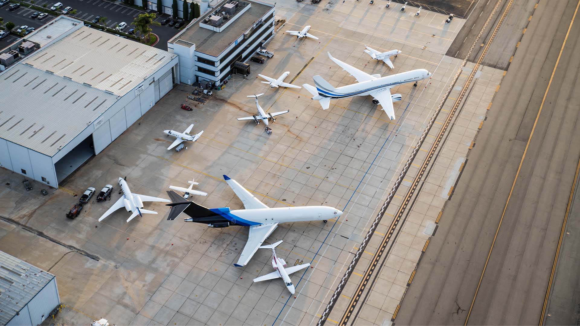Aerial view of several private jets and commercial airplanes parked on an airport tarmac next to hangars.