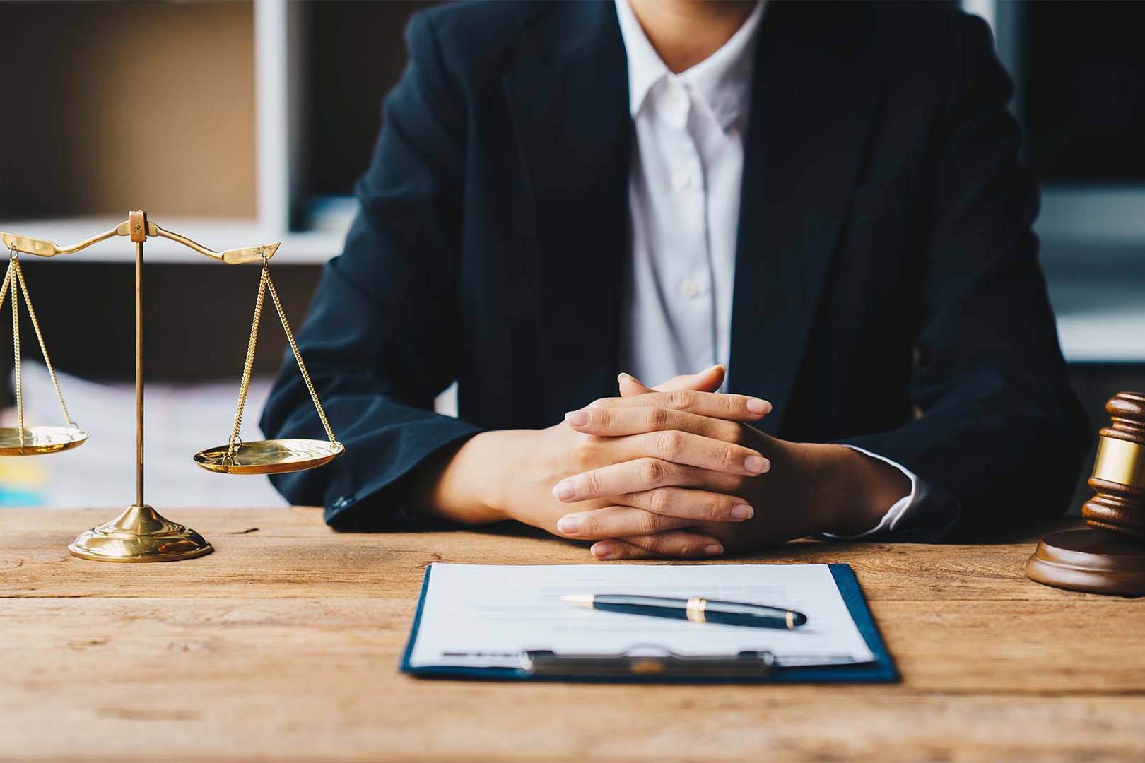 Close-up of a legal professional's hands clasped on a wooden desk with scales of justice and a gavel.