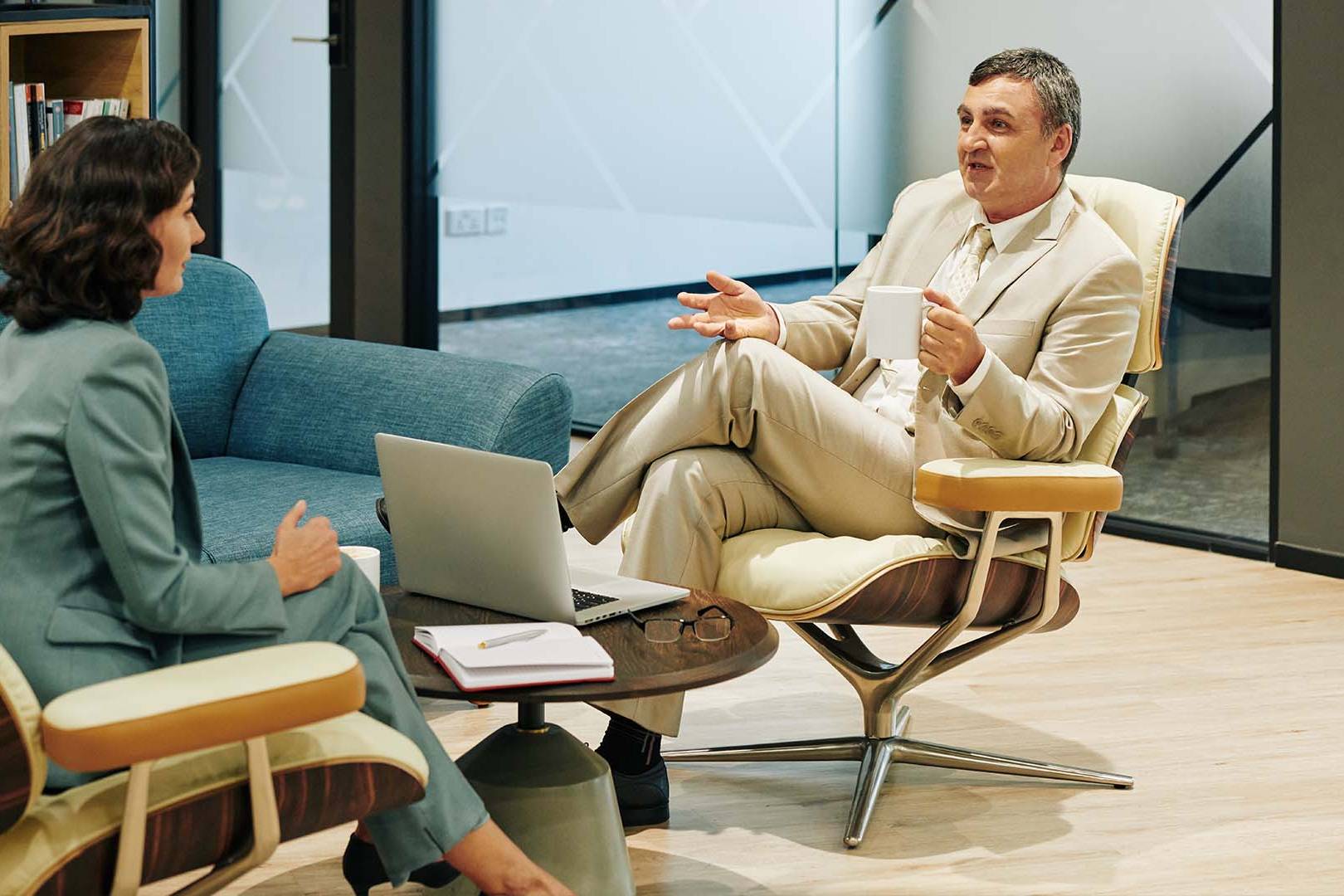 Male executive in a light tan suit talking with a female therapist or consultant in a modern office setting.
