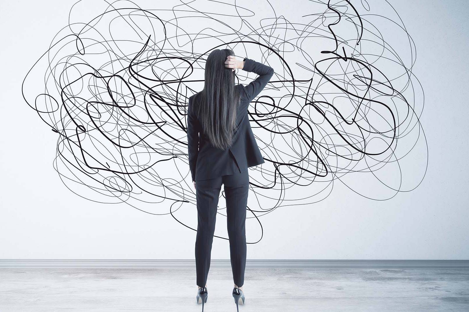 A businesswoman in a dark suit facing a wall covered in a chaotic tangle of black lines.