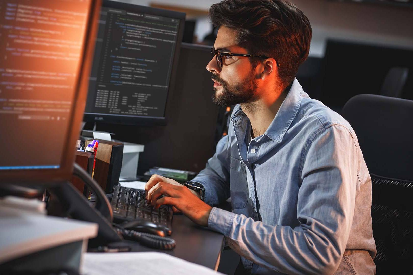 Focused male e-commerce founder with glasses working late at night on dual computer monitors.