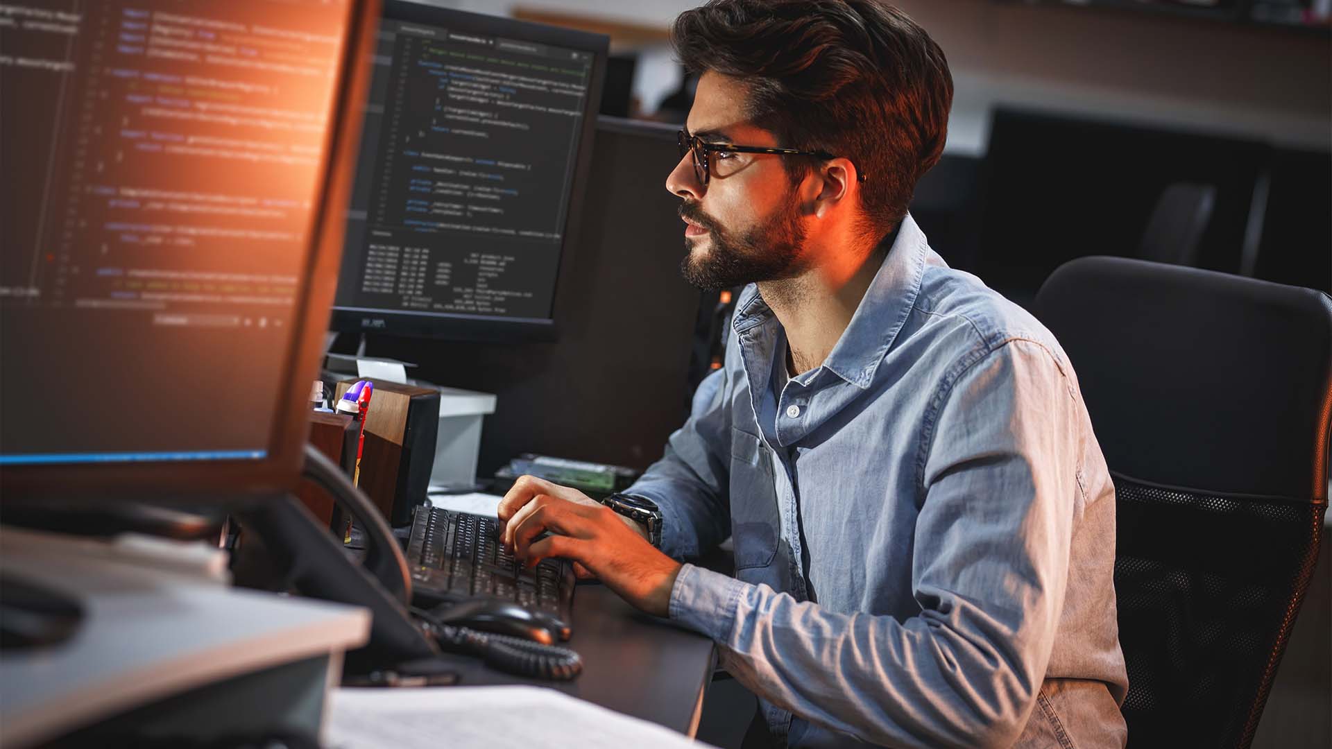 Focused male e-commerce founder with glasses working late at night on dual computer monitors.