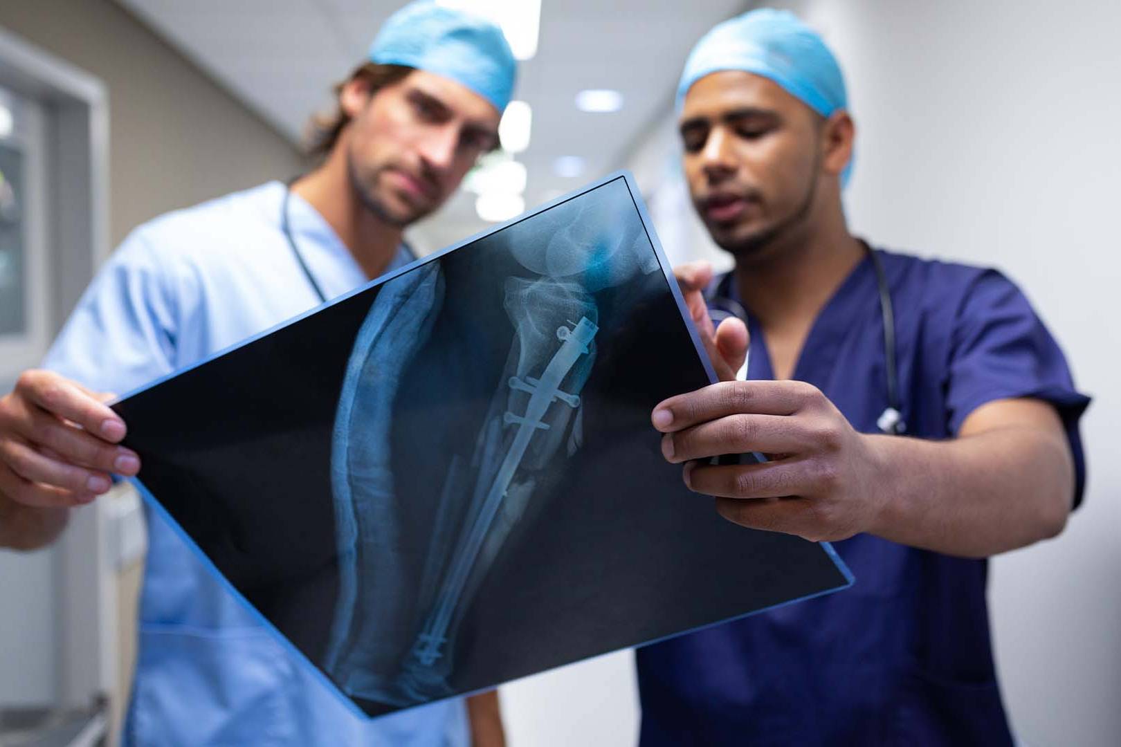 Two male orthopedic surgeons in blue scrubs looking closely at a leg x-ray.