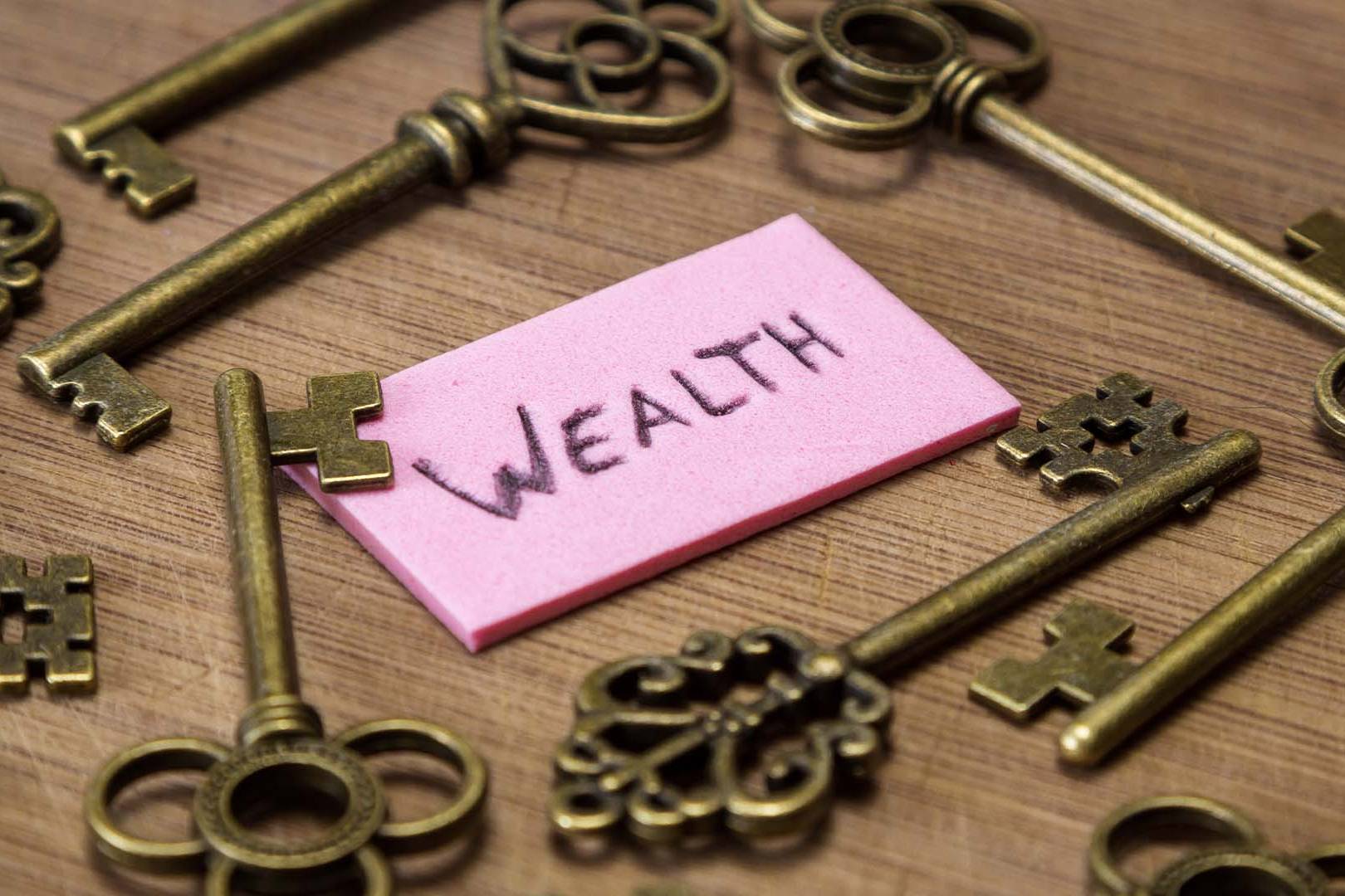 A pink sticky note with the word WEALTH surrounded by vintage brass keys on a wooden table.