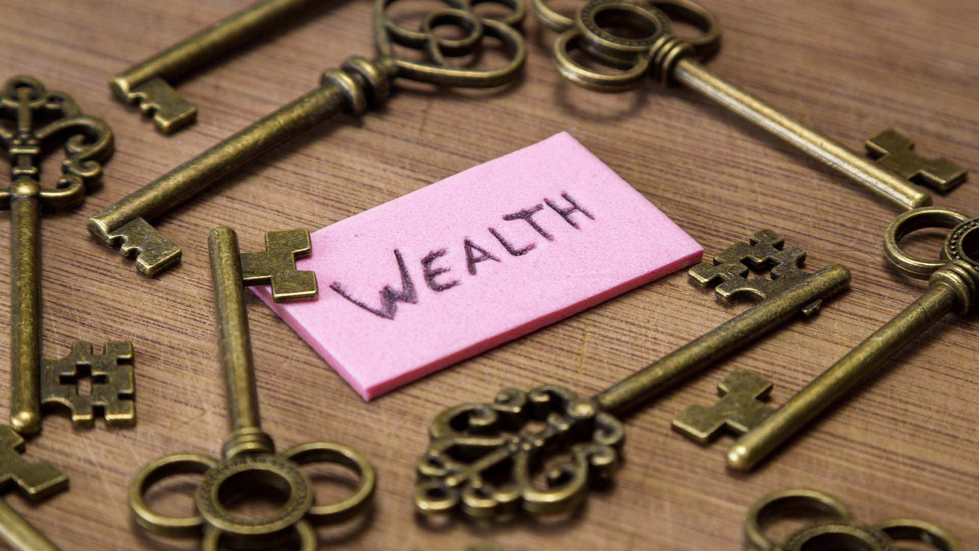 A pink sticky note with the word WEALTH surrounded by vintage brass keys on a wooden table.