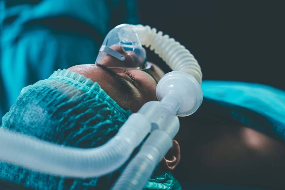 A patient lying on an operating table wearing a surgical cap and an anesthesia mask connected to breathing tubes, under cool blue-green lighting in an operating room.