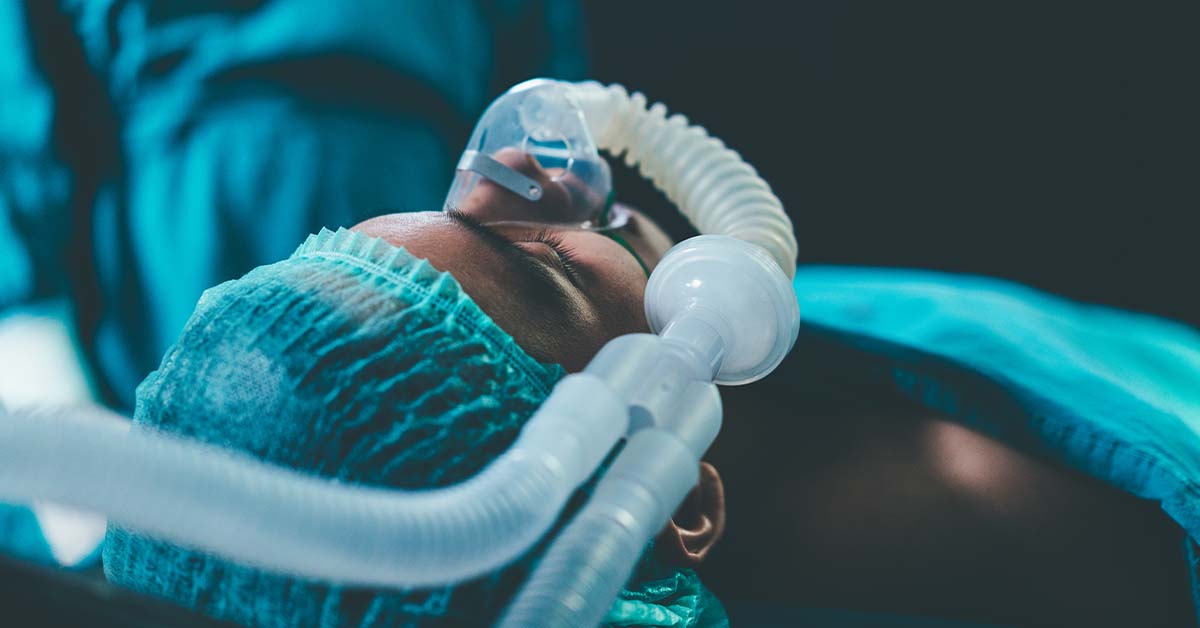 A patient lying on an operating table wearing a surgical cap and an anesthesia mask connected to breathing tubes, under cool blue-green lighting in an operating room.