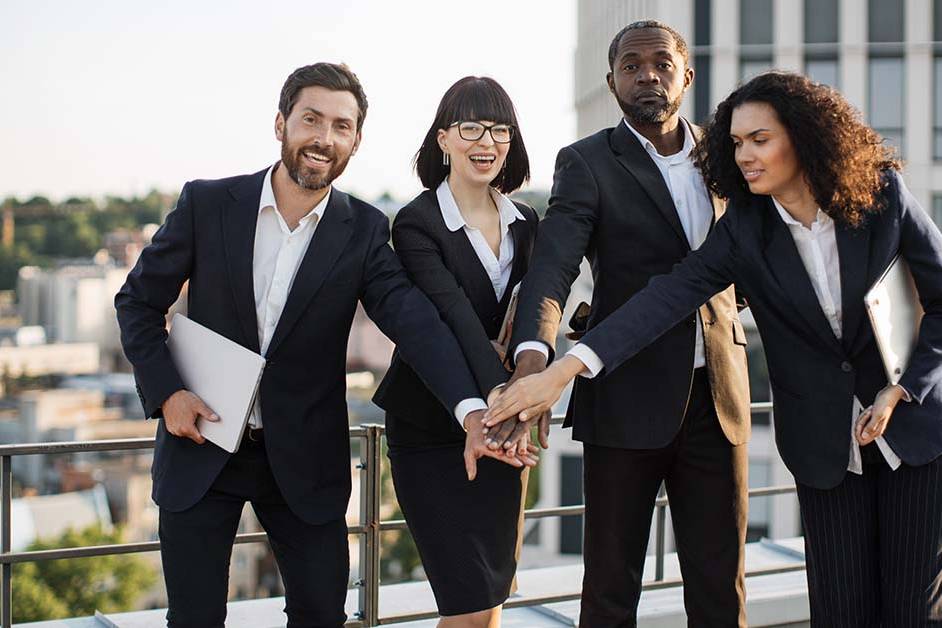 Four diverse business professionals in suits stack their hands together in a team gesture on a rooftop terrace overlooking a city, smiling with a sense of unity and success.