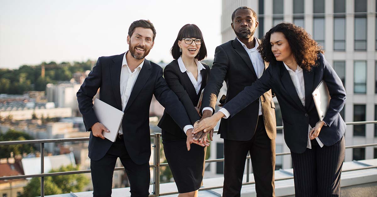 Four diverse business professionals in suits stack their hands together in a team gesture on a rooftop terrace overlooking a city, smiling with a sense of unity and success.
