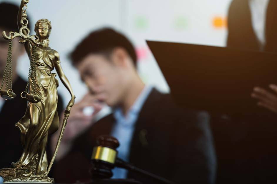 A golden Lady Justice statue holding scales stands in sharp focus beside a gavel, with blurred lawyers conferring over documents in the background, representing the legal profession.