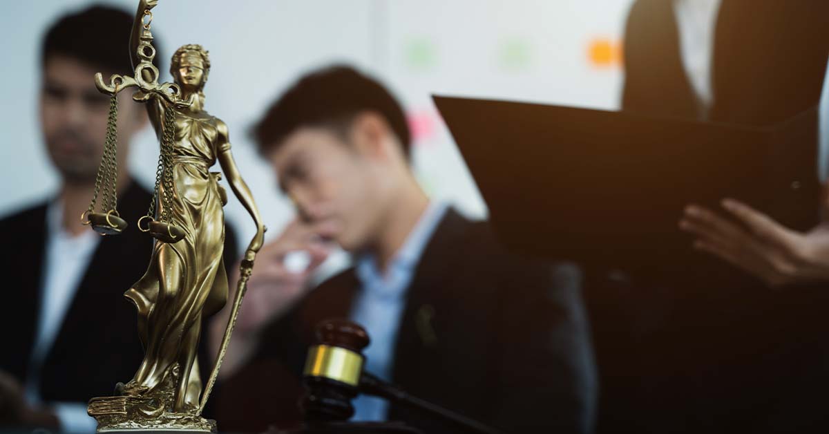 A golden Lady Justice statue holding scales stands in sharp focus beside a gavel, with blurred lawyers conferring over documents in the background, representing the legal profession.