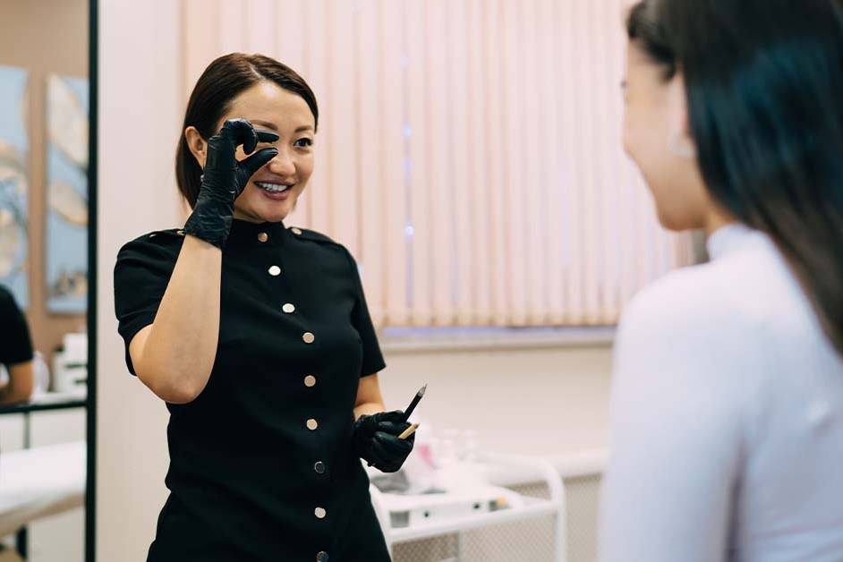 A smiling aesthetician in a black uniform and gloves holds up a microblading or cosmetic tool and looks toward a female client in a bright beauty clinic, appearing to assess or discuss a treatment.