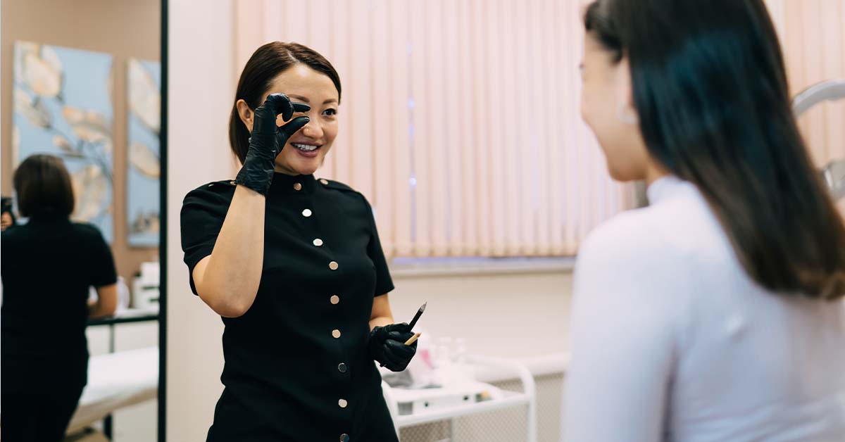 A smiling aesthetician in a black uniform and gloves holds up a microblading or cosmetic tool and looks toward a female client in a bright beauty clinic, appearing to assess or discuss a treatment.
