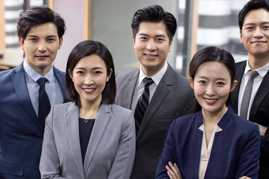 A group of five smiling young Asian business professionals in suits stand together confidently in a modern office, representing the team-driven culture of a Big 4 accounting or consulting firm.