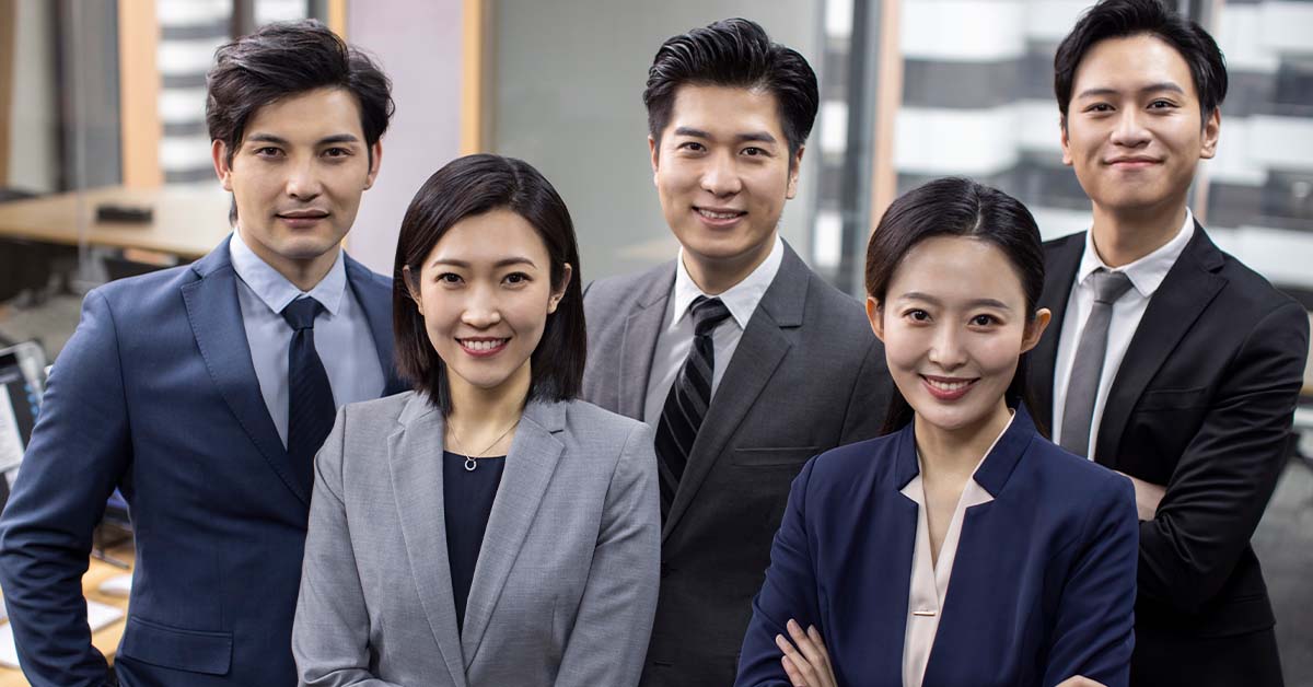 A group of five smiling young Asian business professionals in suits stand together confidently in a modern office, representing the team-driven culture of a Big 4 accounting or consulting firm.