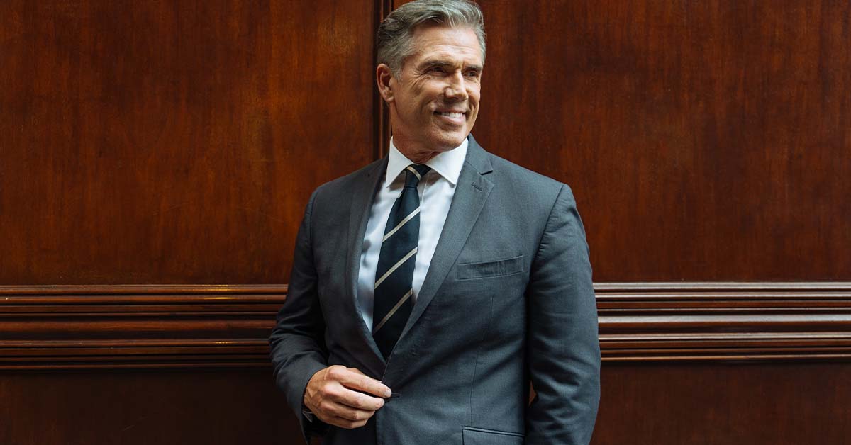 A smiling male attorney in a grey suit standing against a wood-paneled wall, featured image for the article Confidential Therapy for Chicago Attorneys.