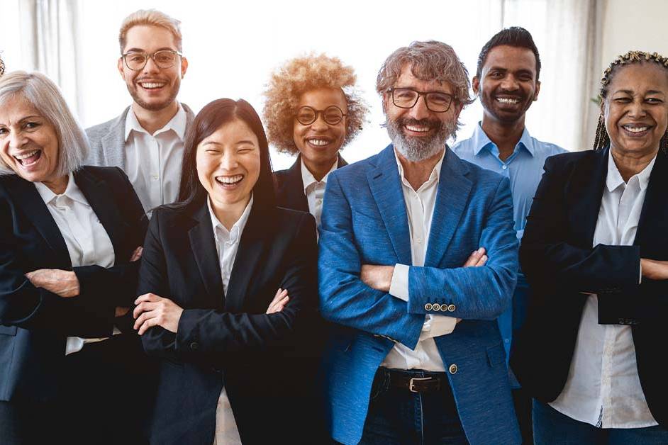A diverse team of five business professionals in smart business-casual attire stand together outdoors in front of modern glass skyscrapers, looking confidently at the camera on a sunny day.