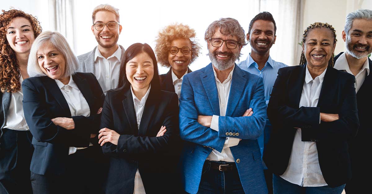 A diverse team of five business professionals in smart business-casual attire stand together outdoors in front of modern glass skyscrapers, looking confidently at the camera on a sunny day.
