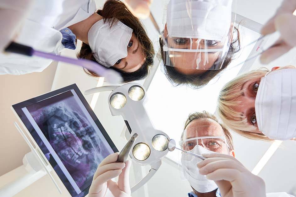 Patient's-eye view looking up at a dental team of four masked professionals wearing white coats, face shields, and gloves, gathered around an overhead dental light and holding instruments, with a dental X-ray displayed on a screen to the left.