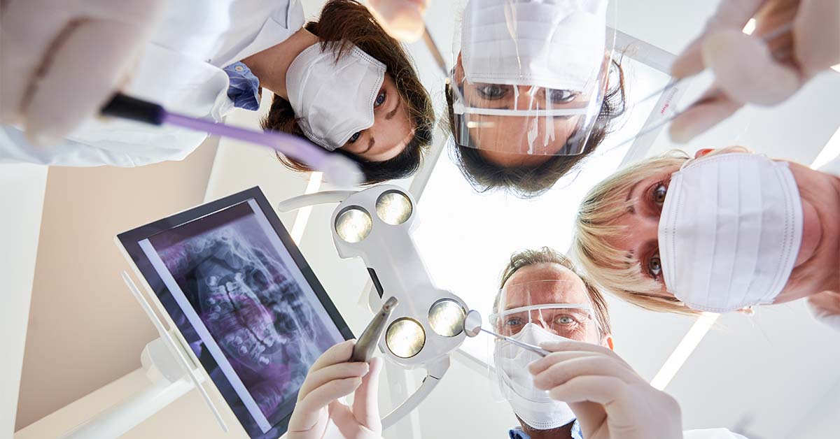 Patient's-eye view looking up at a dental team of four masked professionals wearing white coats, face shields, and gloves, gathered around an overhead dental light and holding instruments, with a dental X-ray displayed on a screen to the left.