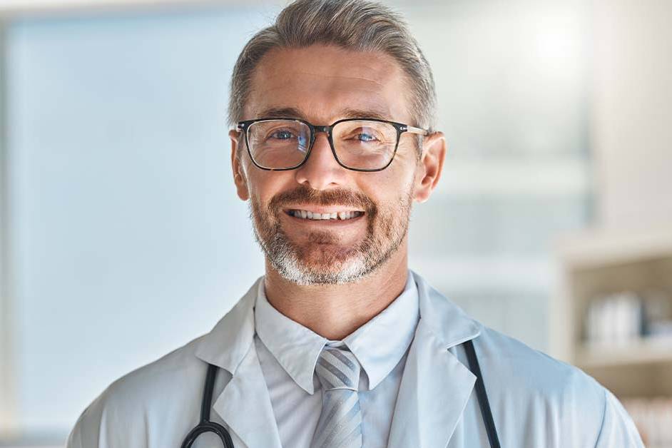 A smiling middle-aged male doctor with gray hair, a beard, and glasses, wearing a white coat and stethoscope, looking confidently at the camera in a bright clinic.