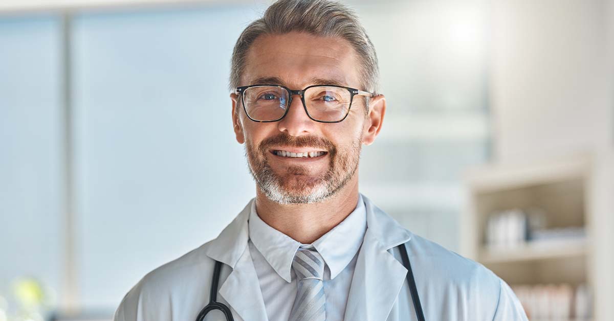 A smiling middle-aged male doctor with gray hair, a beard, and glasses, wearing a white coat and stethoscope, looking confidently at the camera in a bright clinic.