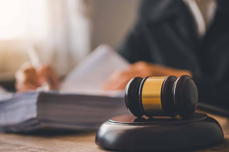 A wooden judge's gavel resting on a sound block in the foreground, with a blurred lawyer or judge in a dark suit writing on documents and a stack of legal papers in the background.