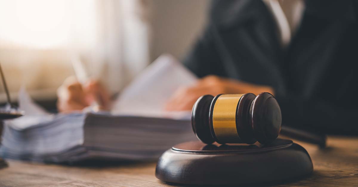 A wooden judge's gavel resting on a sound block in the foreground, with a blurred lawyer or judge in a dark suit writing on documents and a stack of legal papers in the background.