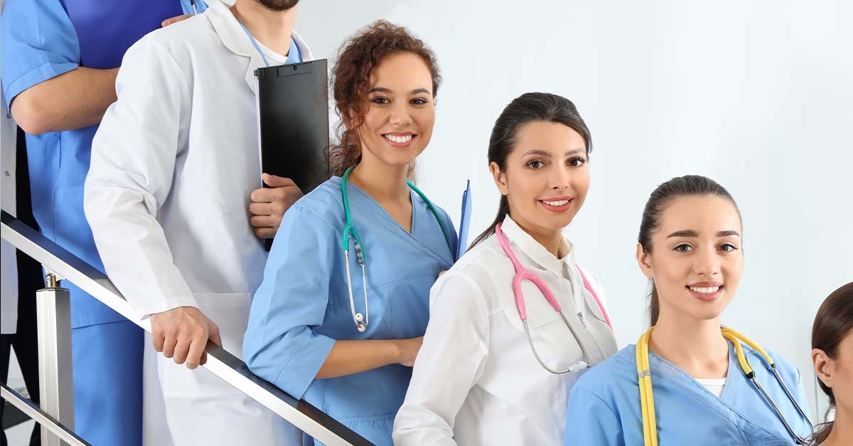 A group of smiling young physicians and medical students in white coats and blue scrubs with colorful stethoscopes stand together on a staircase, representing a medical society's members.
