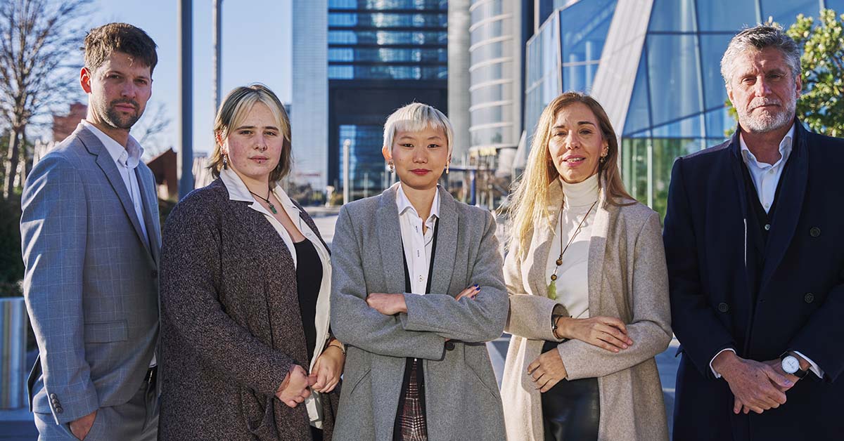 A diverse team of five business professionals in smart business-casual attire stand together outdoors in front of modern glass skyscrapers, looking confidently at the camera on a sunny day.