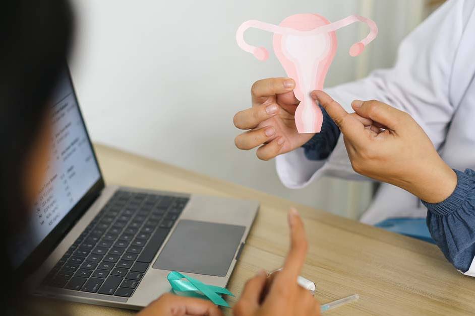 A doctor in a white coat holds up a paper cutout of a uterus and ovaries while consulting with a female patient across a desk, with a laptop and a teal awareness ribbon nearby.