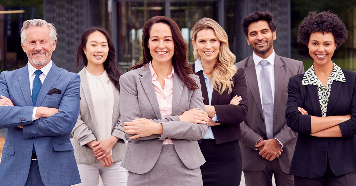 A diverse group of six smiling business professionals in suits stand confidently with arms crossed in front of a modern office building, led by a woman in a gray suit at the center.