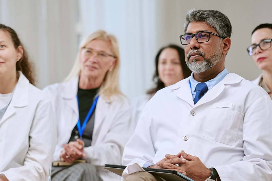 A diverse group of doctors and medical professionals in white coats sit attentively in a row, listening and looking forward as if in a lecture, seminar, or conference setting.