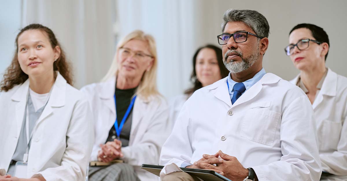 A diverse group of doctors and medical professionals in white coats sit attentively in a row, listening and looking forward as if in a lecture, seminar, or conference setting.