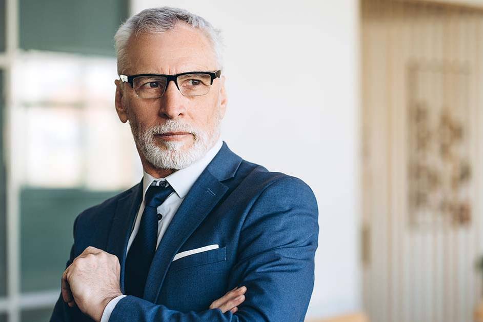 A distinguished senior businessman with grey hair, a beard, and glasses, wearing a navy suit and tie with arms crossed, standing confidently in a modern office—representing a seasoned RIA or wealth manager.
