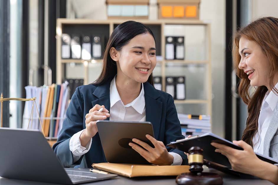 Two smiling businesswomen in suits sit together at a desk reviewing a tablet and documents in a law office, with a brass scales of justice, a gavel, and a laptop on the desk.