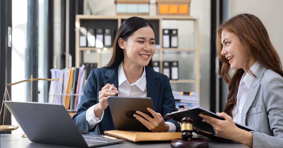 Two smiling businesswomen in suits sit together at a desk reviewing a tablet and documents in a law office, with a brass scales of justice, a gavel, and a laptop on the desk.