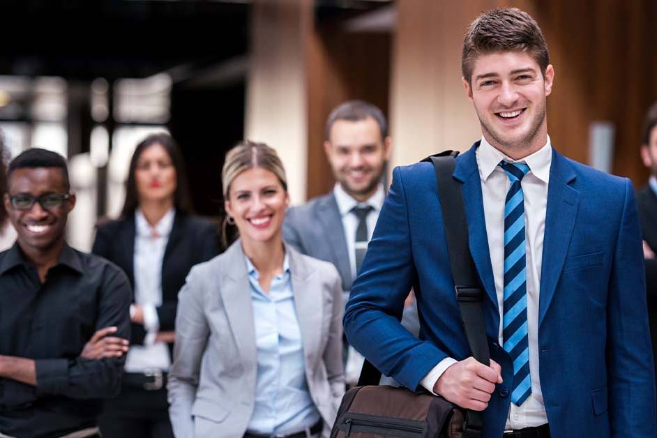 A smiling young businessman with a messenger bag stands in the foreground while a diverse group of confident professionals stands behind him, representing the insured professionals served by liability carriers.