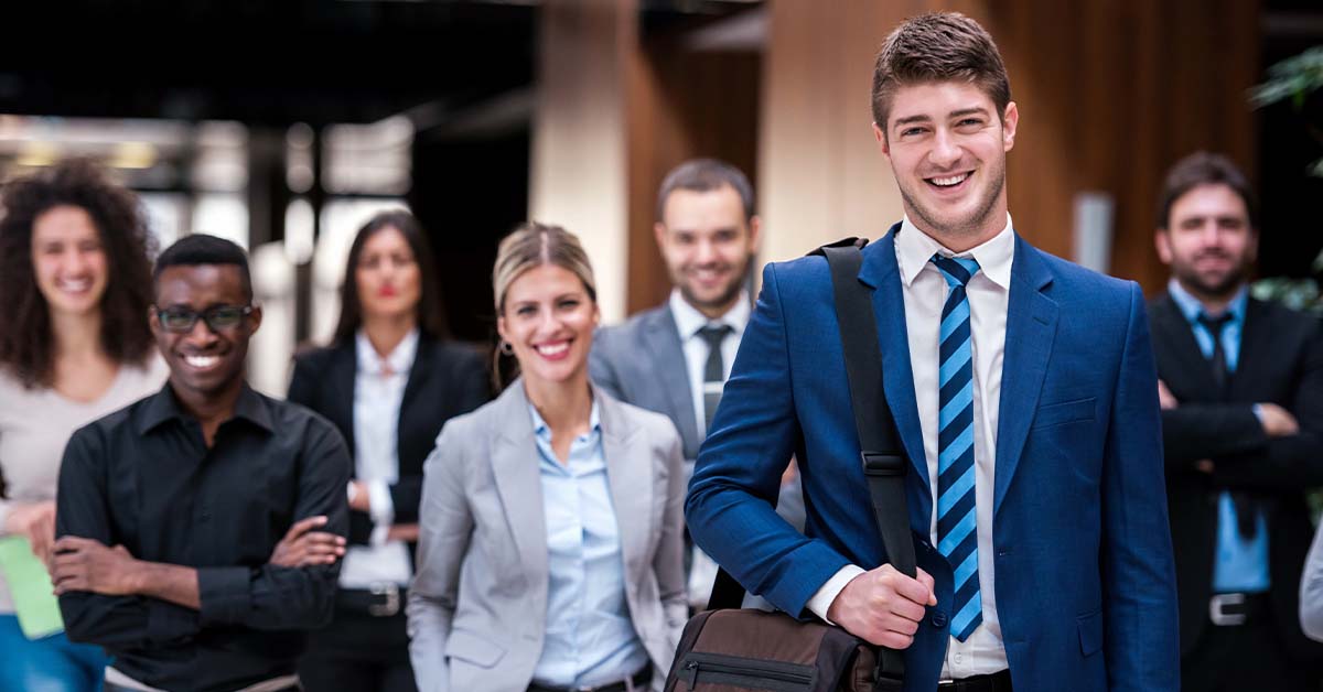 A smiling young businessman with a messenger bag stands in the foreground while a diverse group of confident professionals stands behind him, representing the insured professionals served by liability carriers.