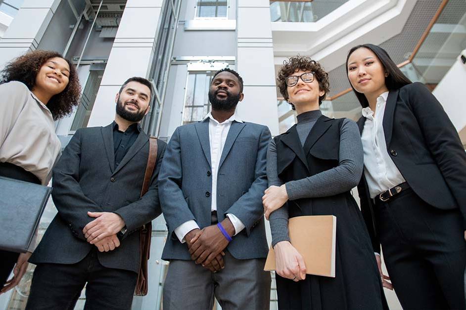 A diverse group of five confident business professionals in suits stand together holding folders and bags, smiling at the camera in a modern office building atrium, shot from a low angle.