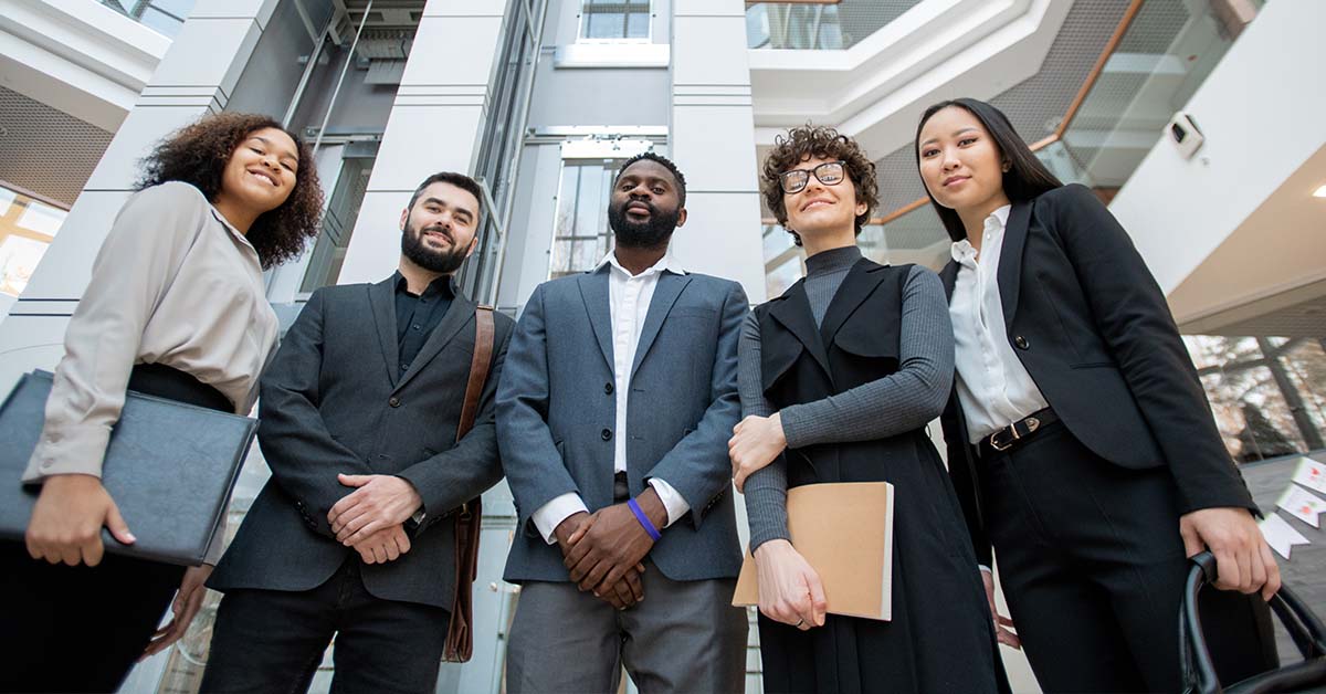A diverse group of five confident business professionals in suits stand together holding folders and bags, smiling at the camera in a modern office building atrium, shot from a low angle.