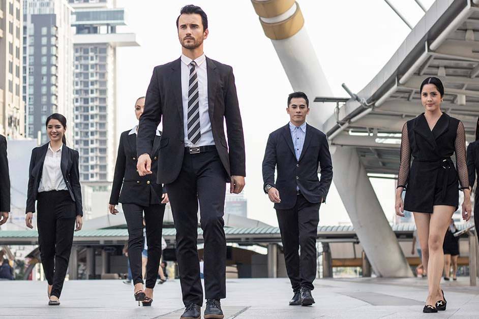 A confident male executive in a dark suit leads a group of business professionals walking forward through a modern urban plaza, representing leadership development and executive coaching.