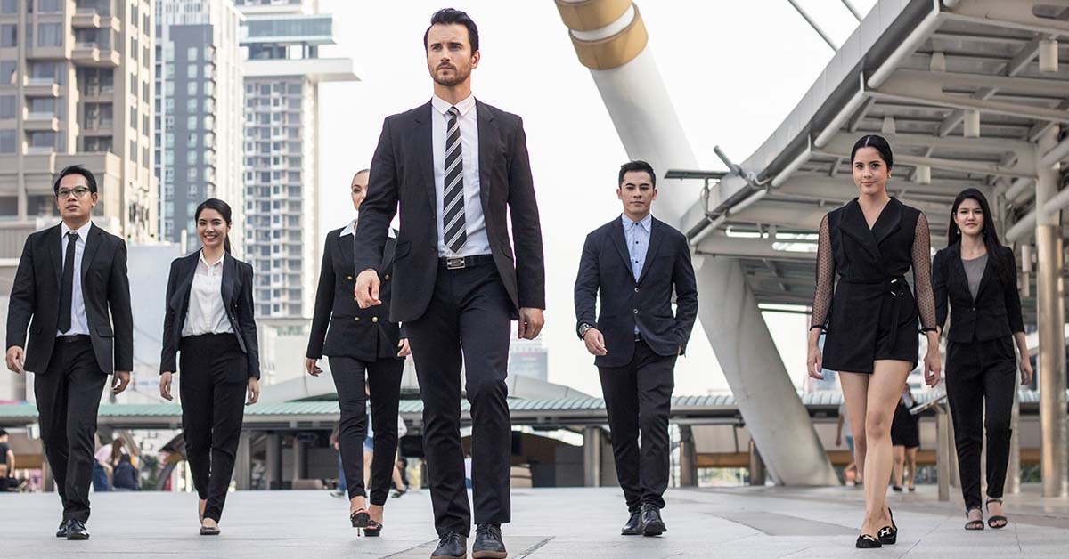 A confident male executive in a dark suit leads a group of business professionals walking forward through a modern urban plaza, representing leadership development and executive coaching.