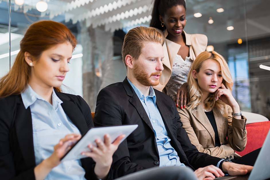 Four diverse business professionals collaborate closely on a couch in a modern office, reviewing a tablet and laptop together, representing the consultative work of a retained search firm.