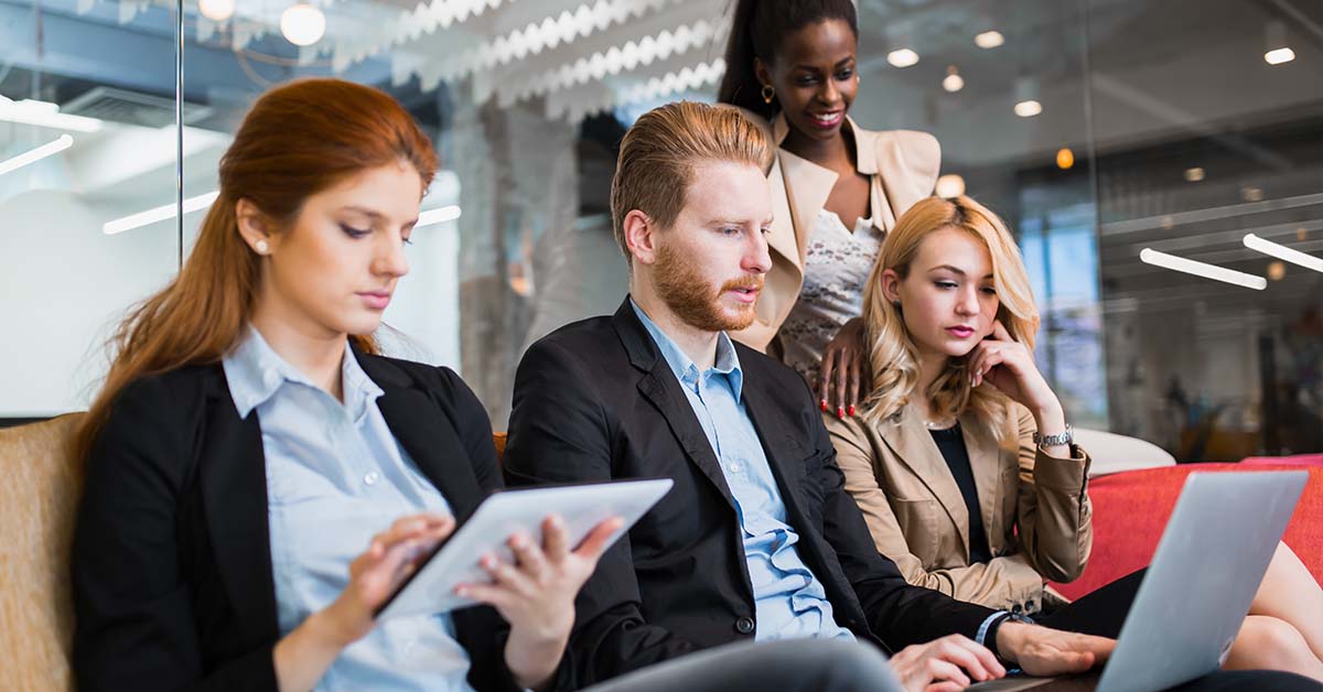 Four diverse business professionals collaborate closely on a couch in a modern office, reviewing a tablet and laptop together, representing the consultative work of a retained search firm.
