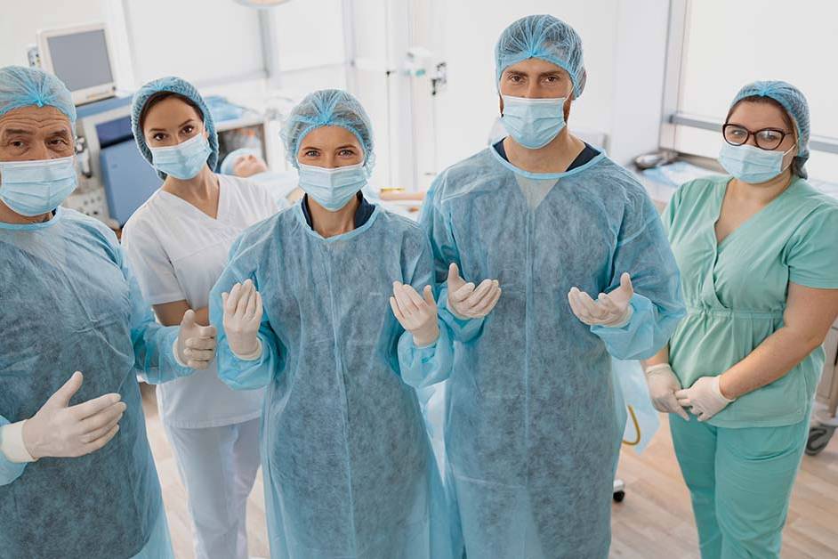 A team of five surgical professionals in scrubs, gowns, caps, masks, and gloves stand together facing the camera in an operating room, with the surgeons holding their gloved hands up ready for surgery.