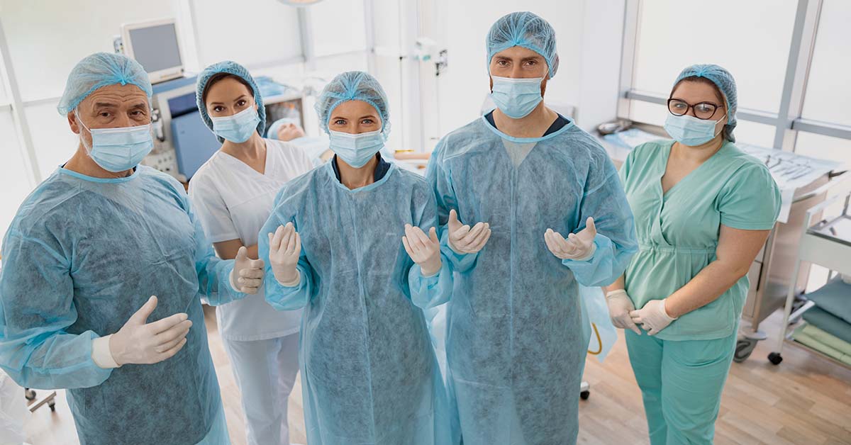 A team of five surgical professionals in scrubs, gowns, caps, masks, and gloves stand together facing the camera in an operating room, with the surgeons holding their gloved hands up ready for surgery.