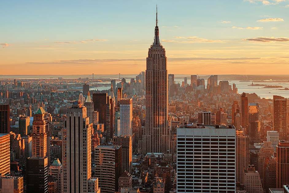 Aerial view of the Manhattan skyline at sunset, with the Empire State Building rising at the center above dense skyscrapers, bathed in warm golden light with the harbor visible in the distance.